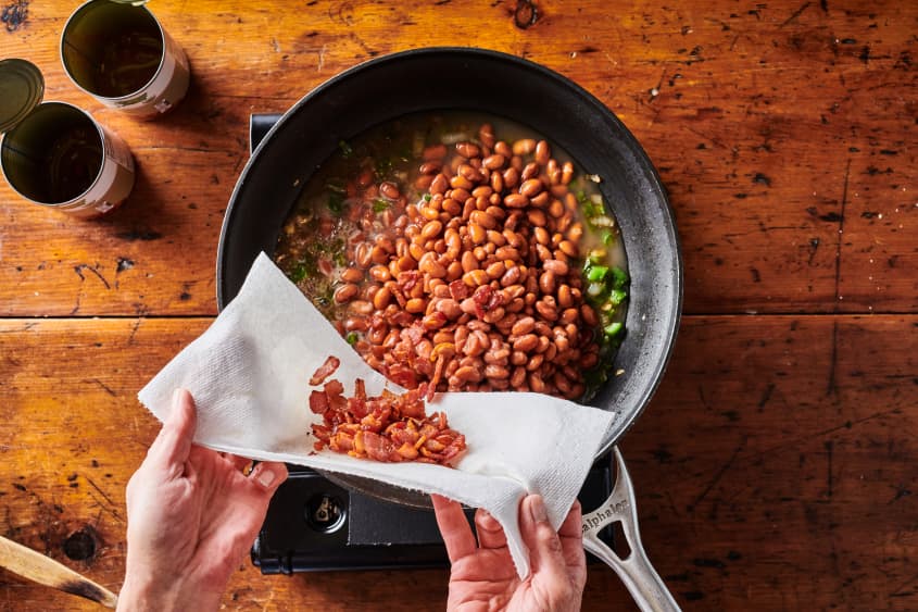 Crisp bacon bits being poured into skillet of beans