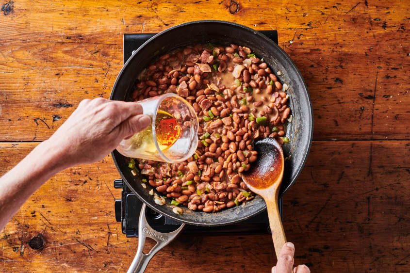 Hand pouring measuring cup of canola oil into skillet cooking with pinto beans, green bell peppers and onions. Right hand stirring beans with wooden spoon