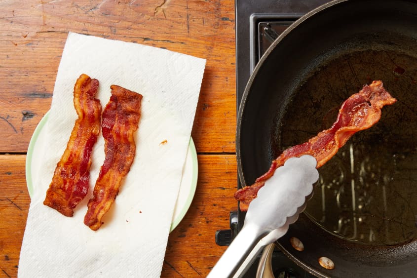 Two crisp pieces of bacon resting on white paper towel. Another piece of bacon being pulled out of skillet with metal tong