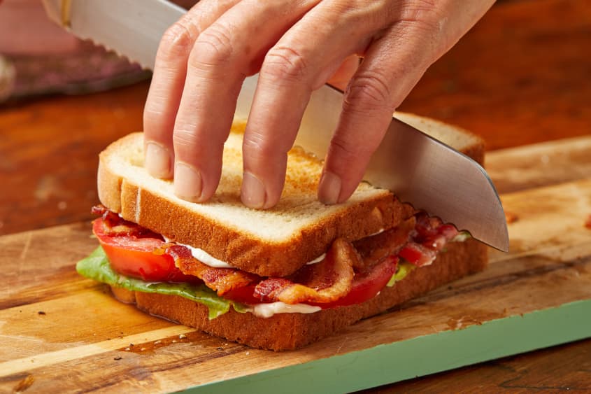 Someone cutting BLT sandwich in half with serrated knife