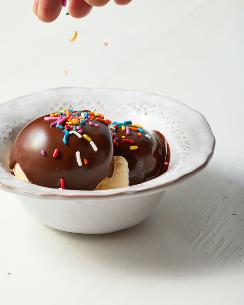 Vintage white bowl filled with two scoops of vanilla ice cream topped with chocolate magic shell and someone adding multi-colored sprinkles