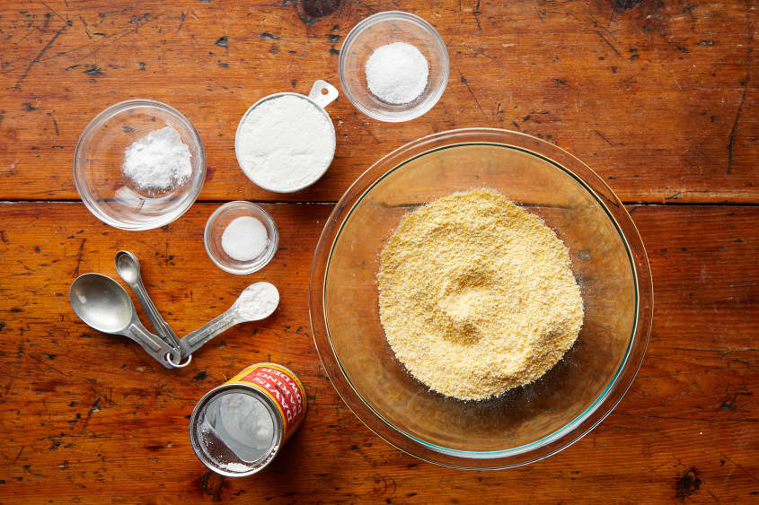 Cornmeal in glass bowl surrounded by dry ingredients: baking powder in tin can, measuring spoon, and various glass bowls filled with sugar, baking soda, salt and AP flour.