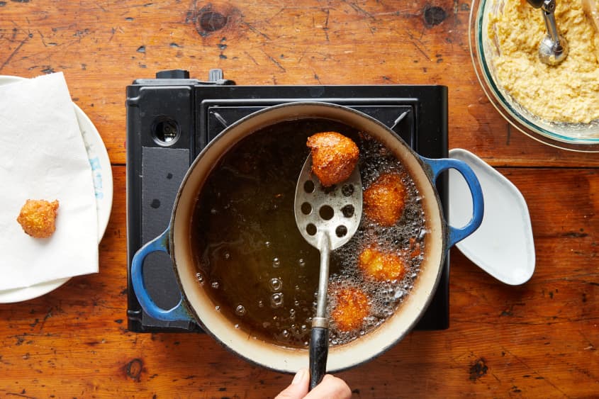 Someone removing fried hushpuppies from blue Dutch oven with slotted spoon. Hushpuppies being transferred to paper towel lined plate.