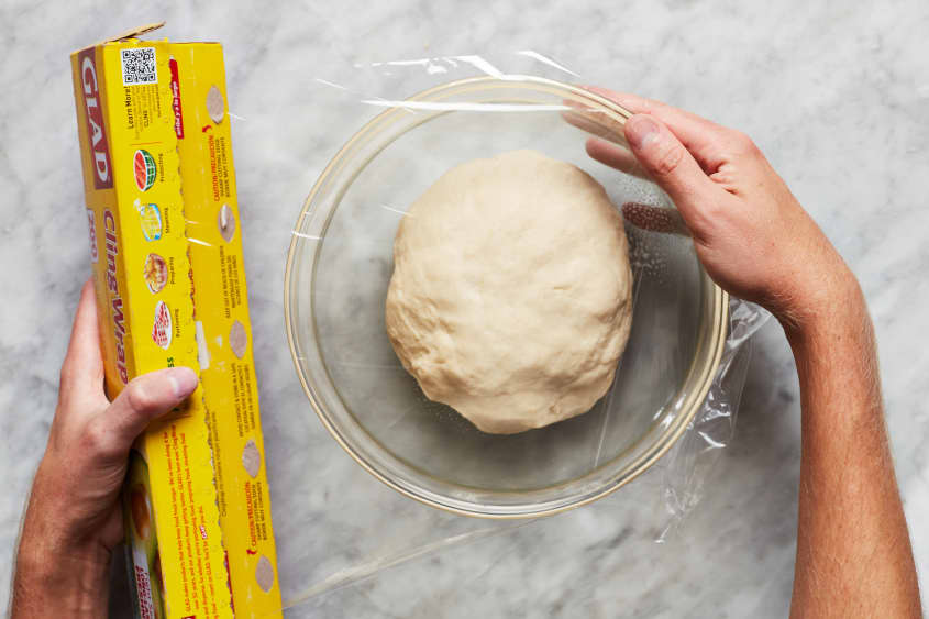 Seran wrap is put over a large glass bowl with dough inside.