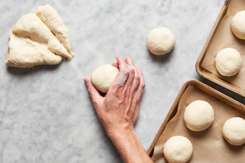 A hand places a ball of dough on a floured surface. Off to the side sit many dough balls on payment lined baking sheets and 3 triangles of dough.