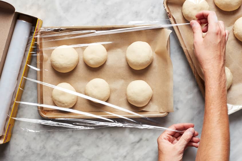 Hands pull serán wrap over six bagel dough balls sitting on a parchment lined baking sheet.