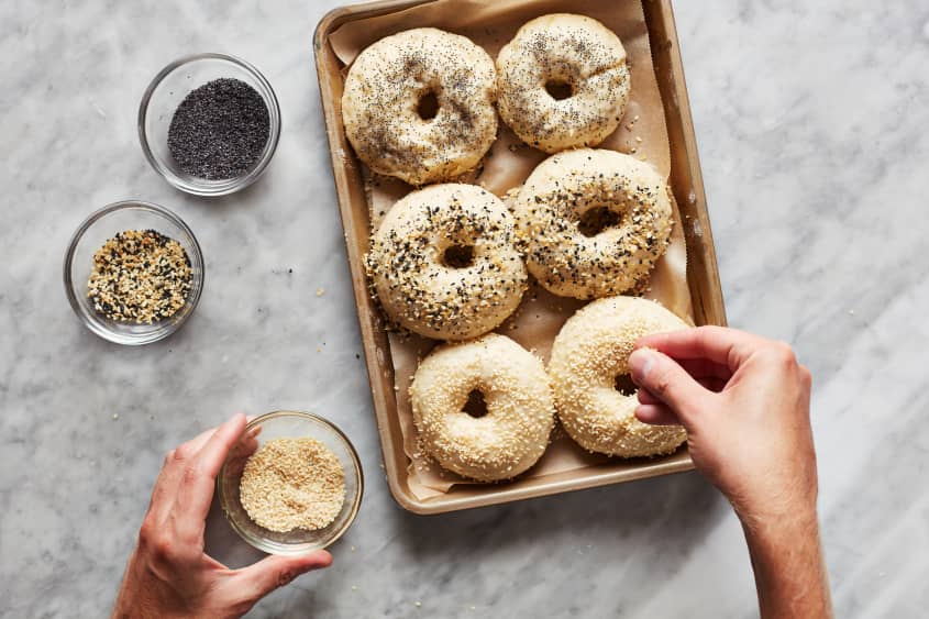 A hand sprinkles everything bagel seasoning over an unbaked bagel, which is nestled into a sheet pan next to five other unbaked bagels.