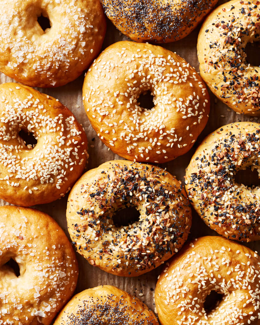 Ten bagels sit on a counter. Each bagel is topped with different seasonings like, everything seasoning, poppyseeds, sesame seeds, and salt.