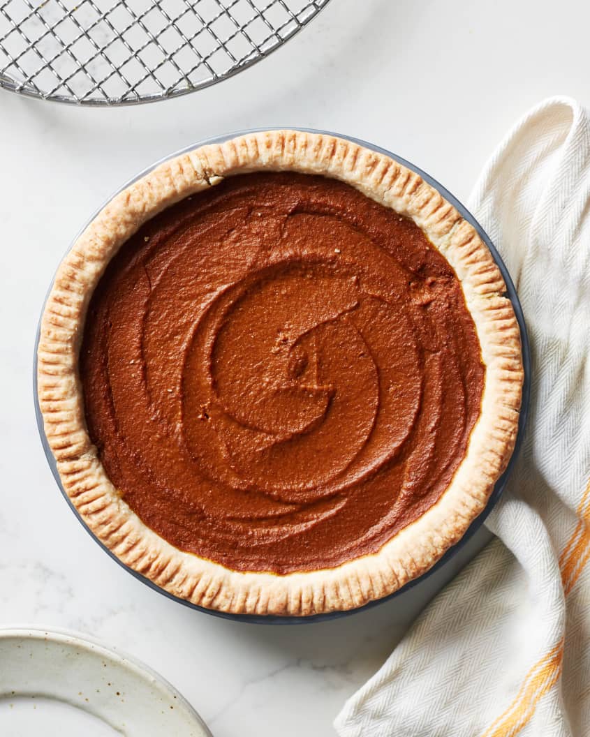 The vegan pumpkin pie is baked and sit on a counter next to the cooling rack, a plate, and a kitchen linen.