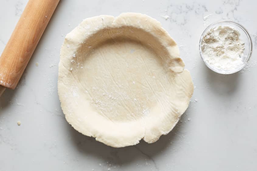 The pie crust as laid over a pie plate, while a bowl of flour and wooden pastry rollers sit on either side of the pie plate.