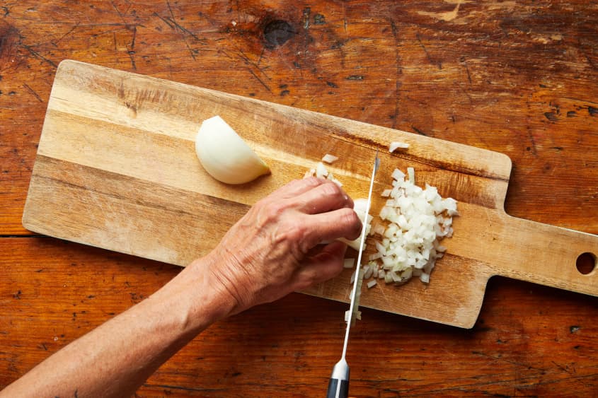 A hand holds an onion against a cutting board as a knife chops the onion.