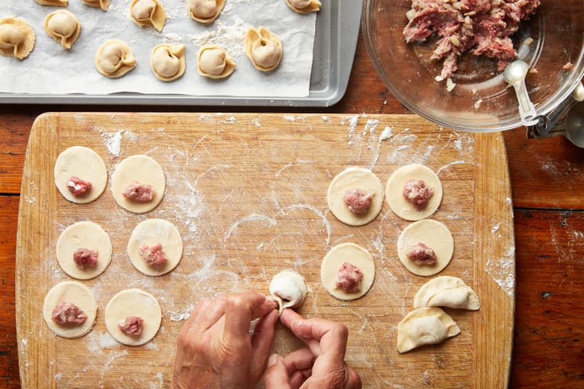 Hands fold a dumpling on a cutting board. Off the side are dough rounds with filling, a sheet pan with folded dumplings, and a bowl of prepared filling.