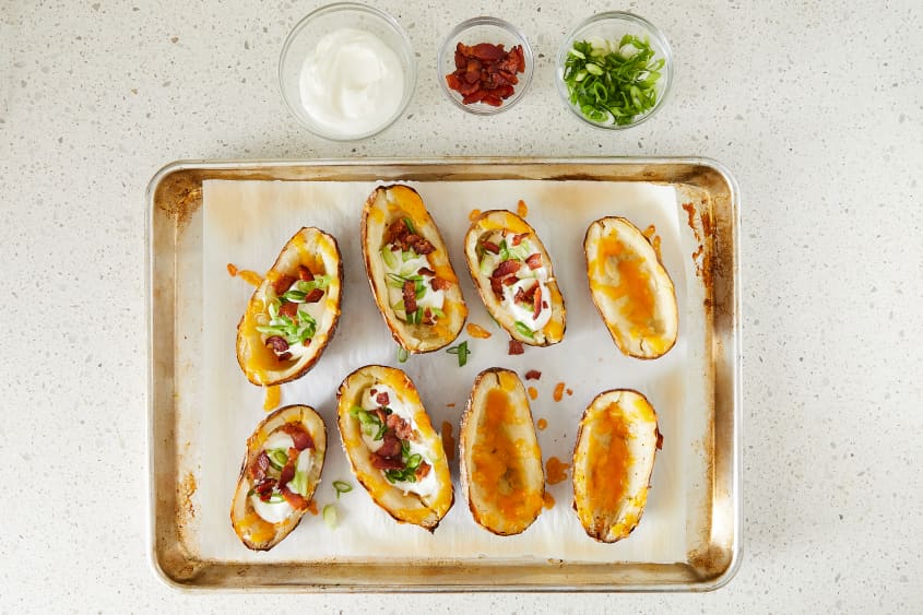 Fully assembled loaded potato skins on baking sheet with a few potatoes missing full toppings. Small glass bowls of sour cream, bacon, and scallions on top of baking sheet.
