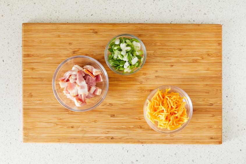 Potato skin toppings in small glass bowls; left to right: cut bacon, diced scallions, shredded cheddar cheese.