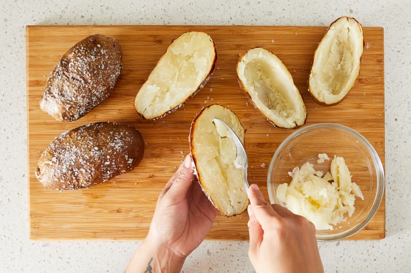 Baked potato halves being scooped out, flesh placed in glass bowl.