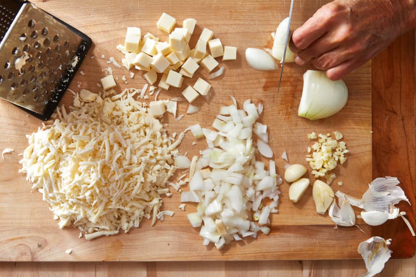 Someone chopping onion on cutting board surrounded by ingredients for cheesy baked rigatoni with beef.