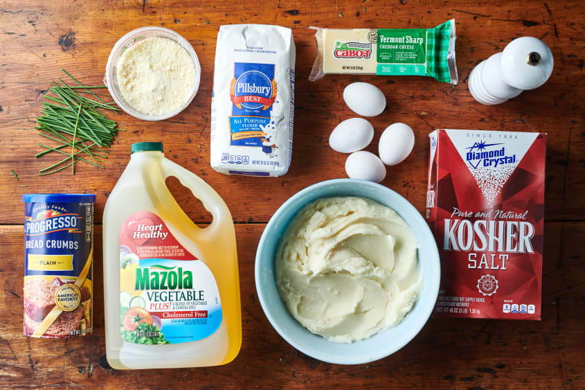 Ingredients laid out for mashed potato croquettes; clockwise: pepper, salt, mashed potatoes, cooking oil, bread crumbs, chives, parmesan, flour, cheddar cheese. Egg in center.