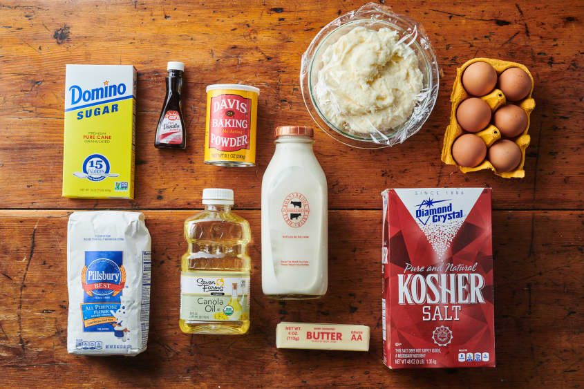 White cake ingredients laid out; clockwise from right: eggs, Kosher salt, butter, milk, canola oil, flour, sugar, vanilla extract, baking powder, buttermilk frosting.