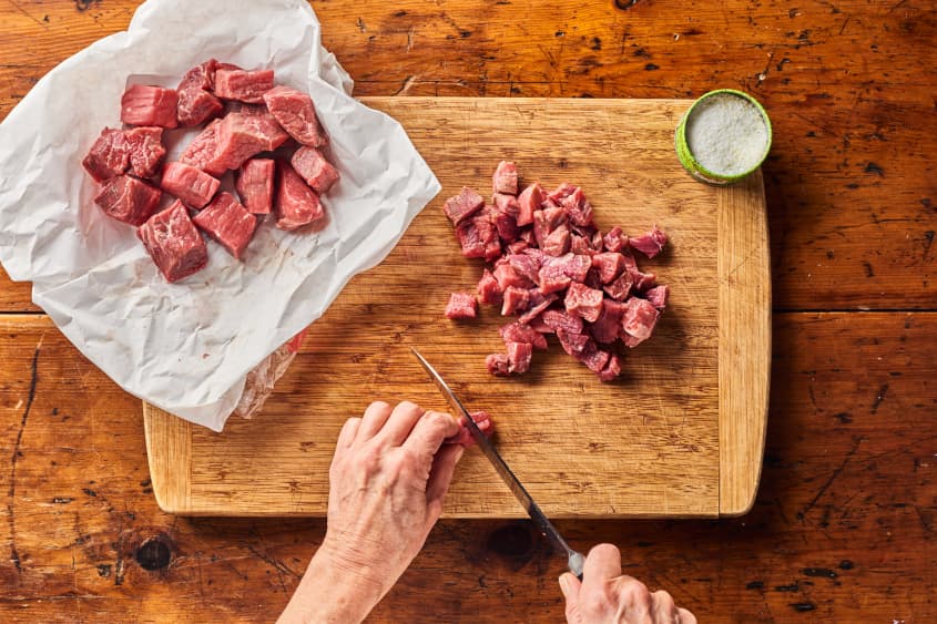 someone is chopping raw beef on a cutting board with larger chunks separated near it in parchment paper