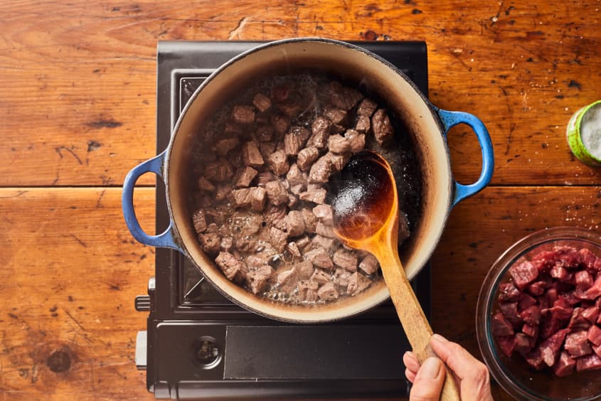 someone is stirring beef chunks with a wooden spoon in a large pot with stock in it