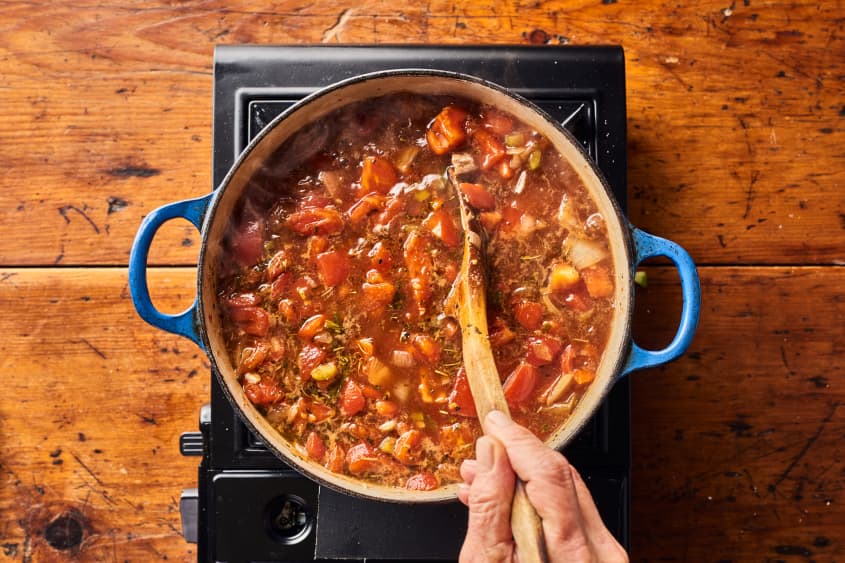 someone is stirring beef, stock, and tomatoes in a large pot with a wooden spoon