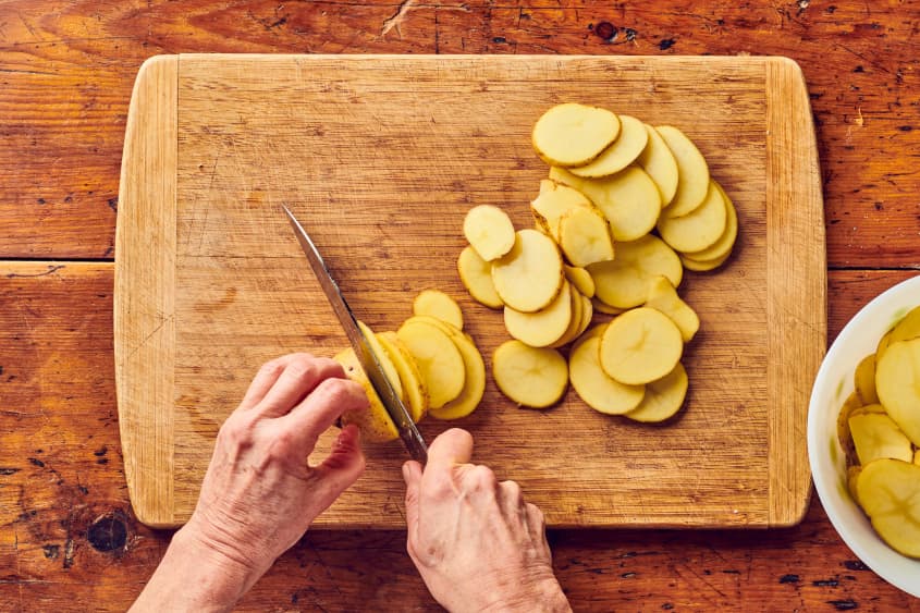 potatoes being sliced by someone into circles on wooden cutting board
