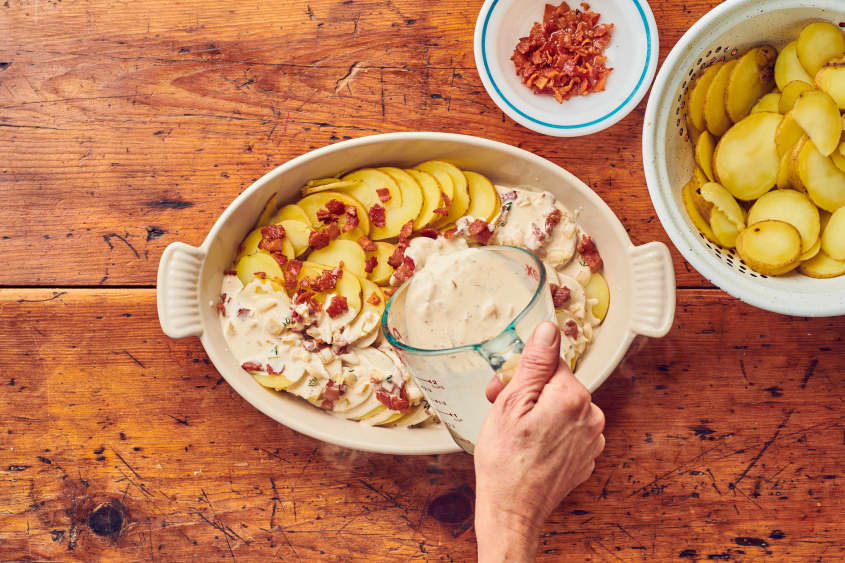 Sauce is being poured over tartiflette where you can see bits of bacon, sliced potatoes, and a small bowl of bacon and more potatoes on the side.
