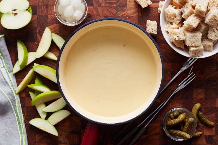 cheese fondue sits on a table next to a bowl of bread, pickles, onions, and sliced apples