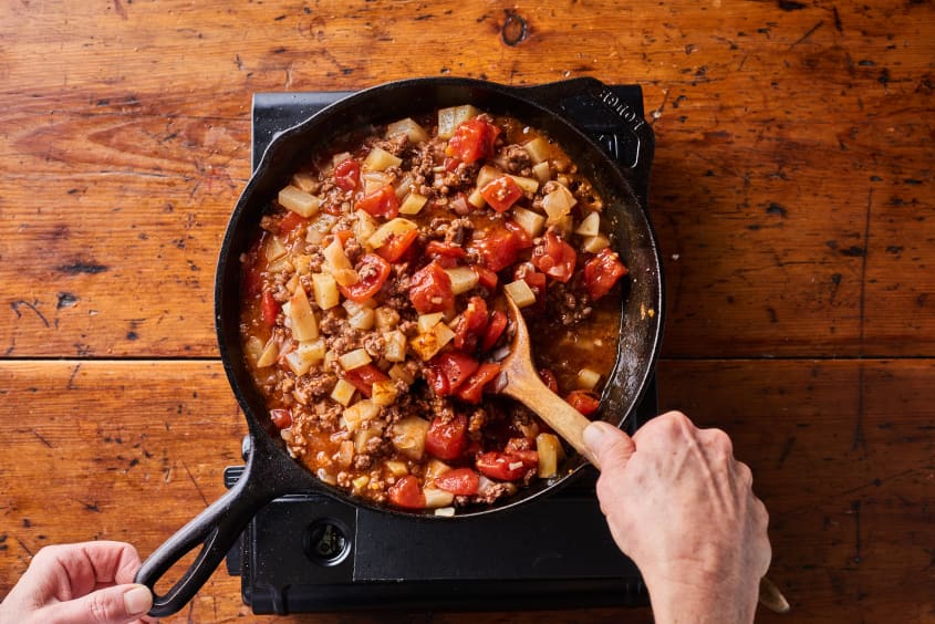 someone is stirring picadillo in a cast iron pot adding tomatoes with a wooden spoon