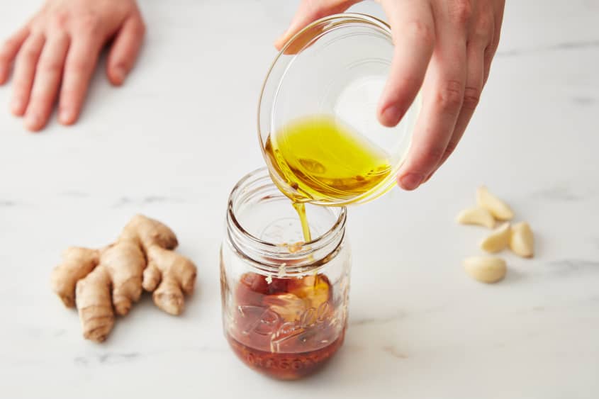 Someone pours olive oil placed in a small dipping bowl into a jar filled with lime juice, tamari or soy sauce, vinegar, honey, ginger, garlic, and chili-garlic paste, with ginger and garlic pictured in the background