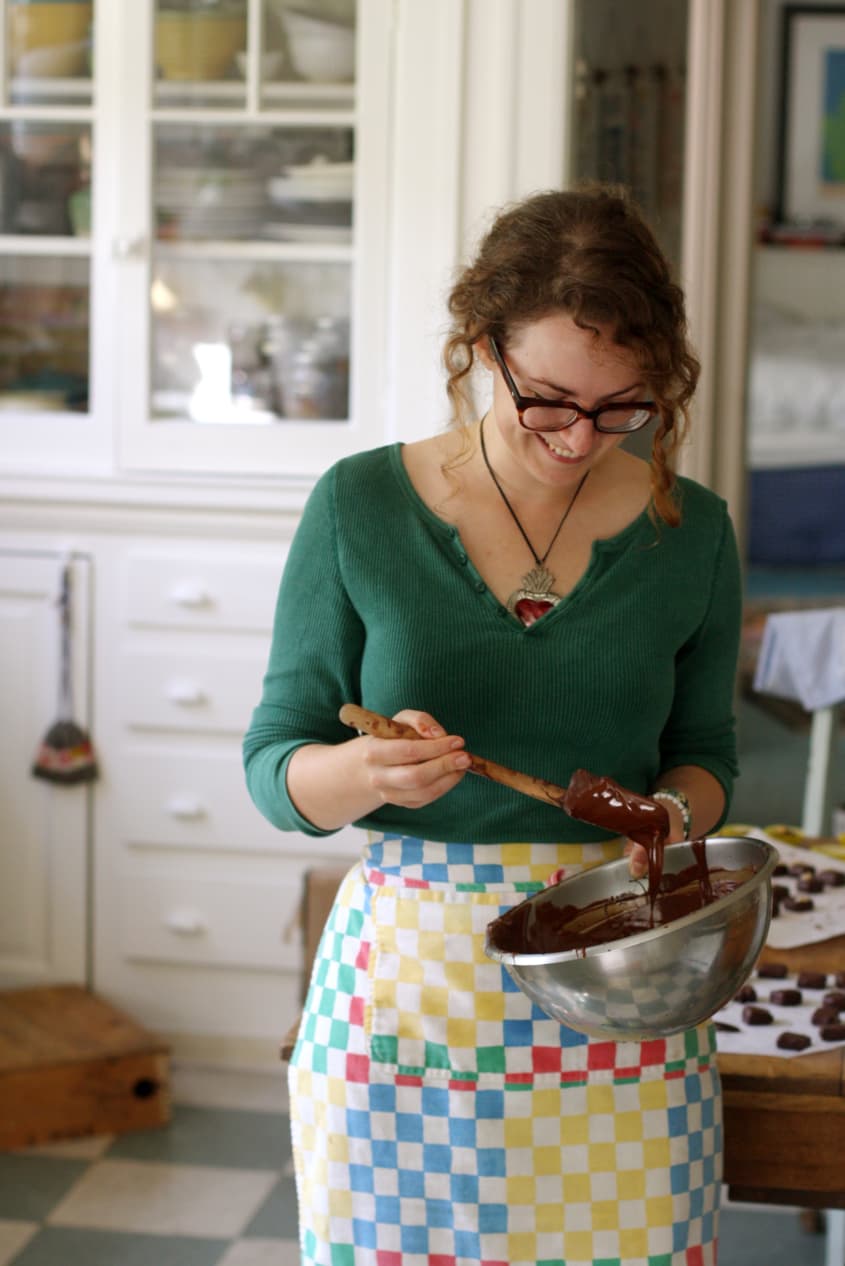 Chocolatier Alexandra Whisnant in long-sleeved green top holds a stainless steel mixing bowl and mixes melted chocolate using a spatula