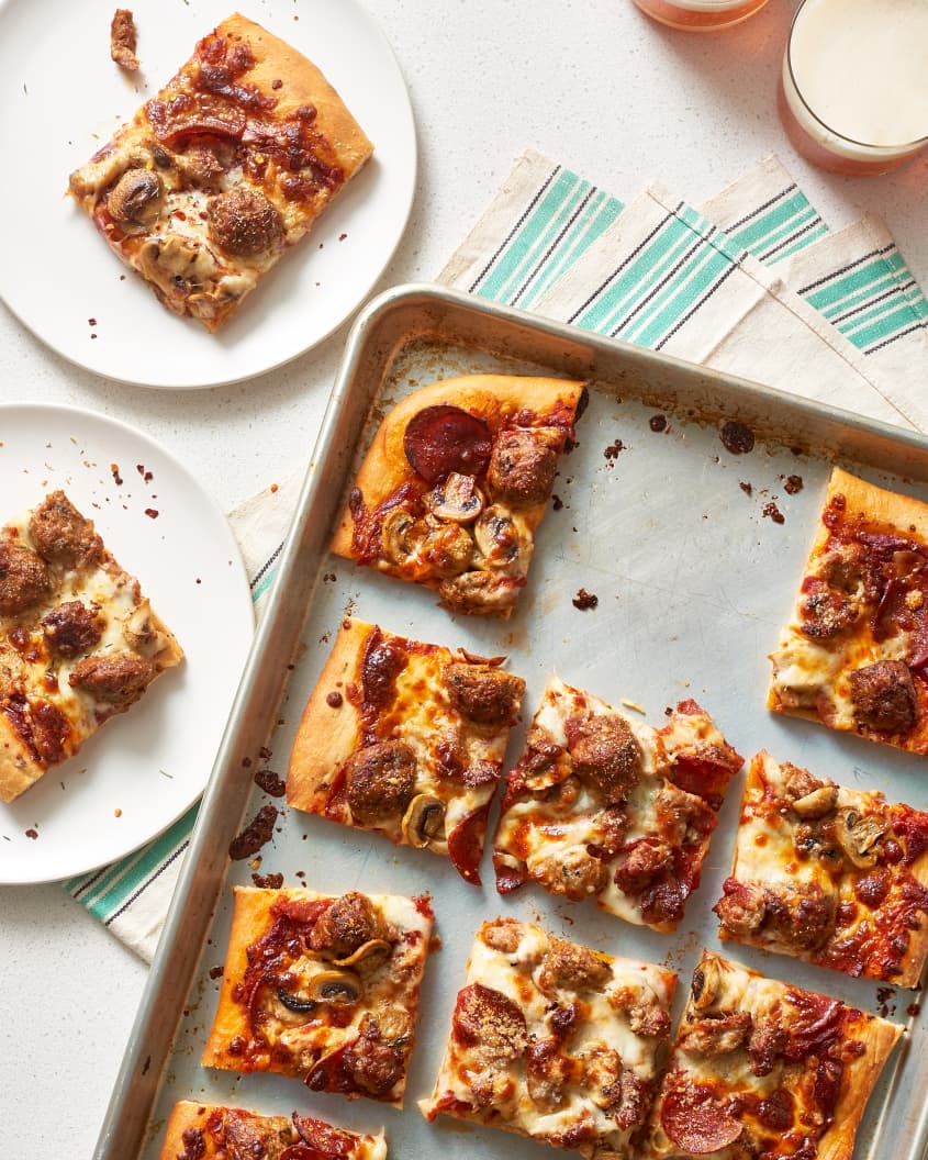 A slice of sheet pan pizza, with tomato sauce, mozzarella cheese, pork sausages, quartered meatball, and pepperoni, on two separate plates and baking sheet