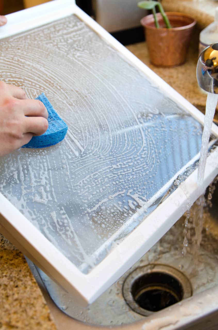 Hand cleaning a soapy window screen with a blue sponge over a kitchen sink.