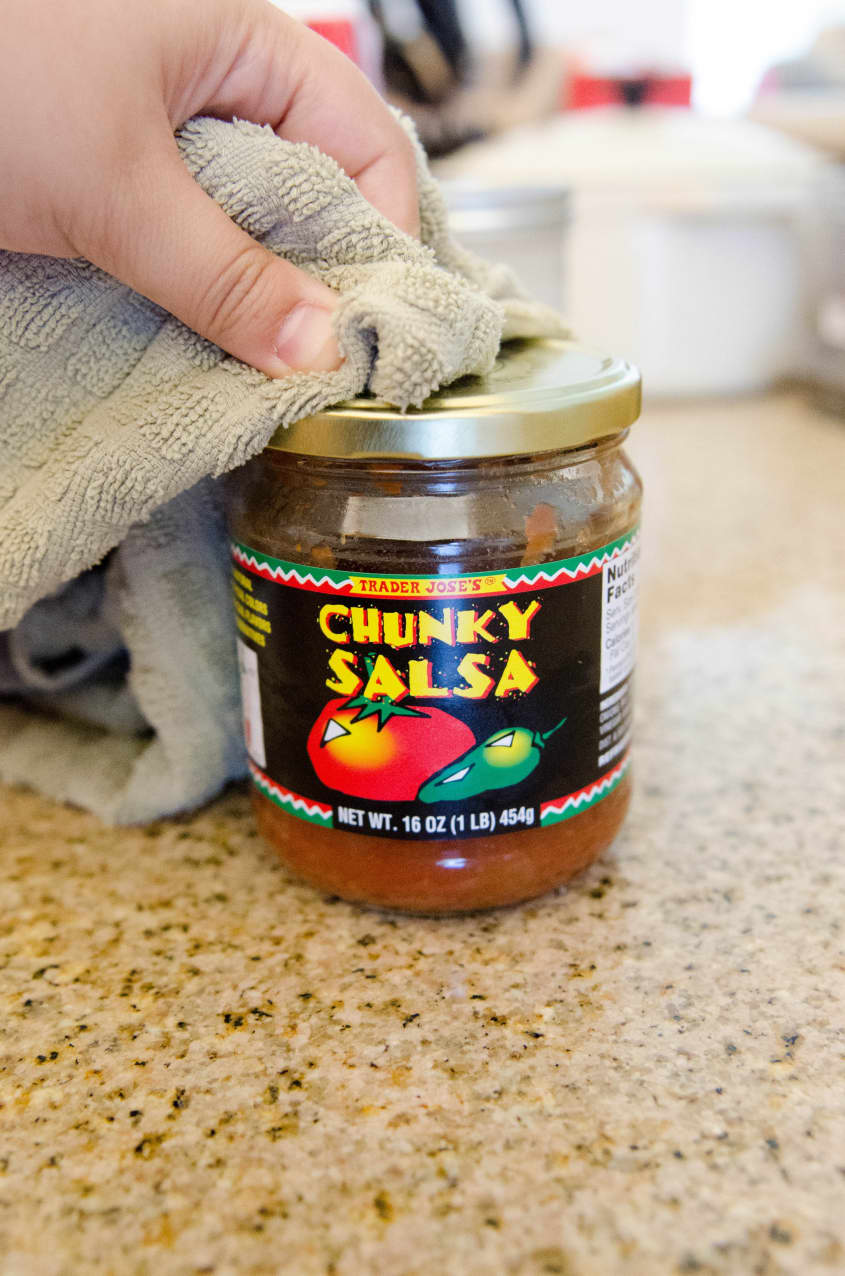 Hand using a cloth to open a jar of Trader Joe's Chunky Salsa on a kitchen counter.