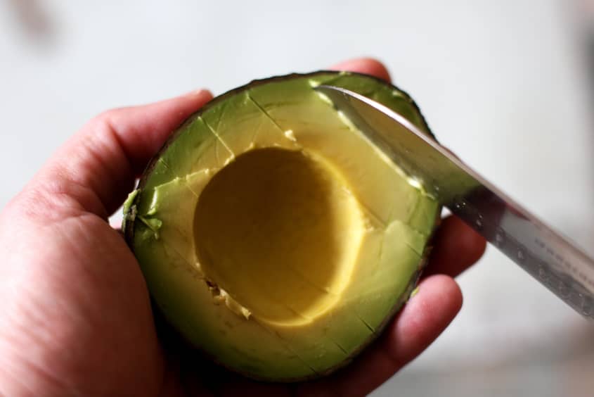 Hand holding half an avocado with a knife slicing the flesh.