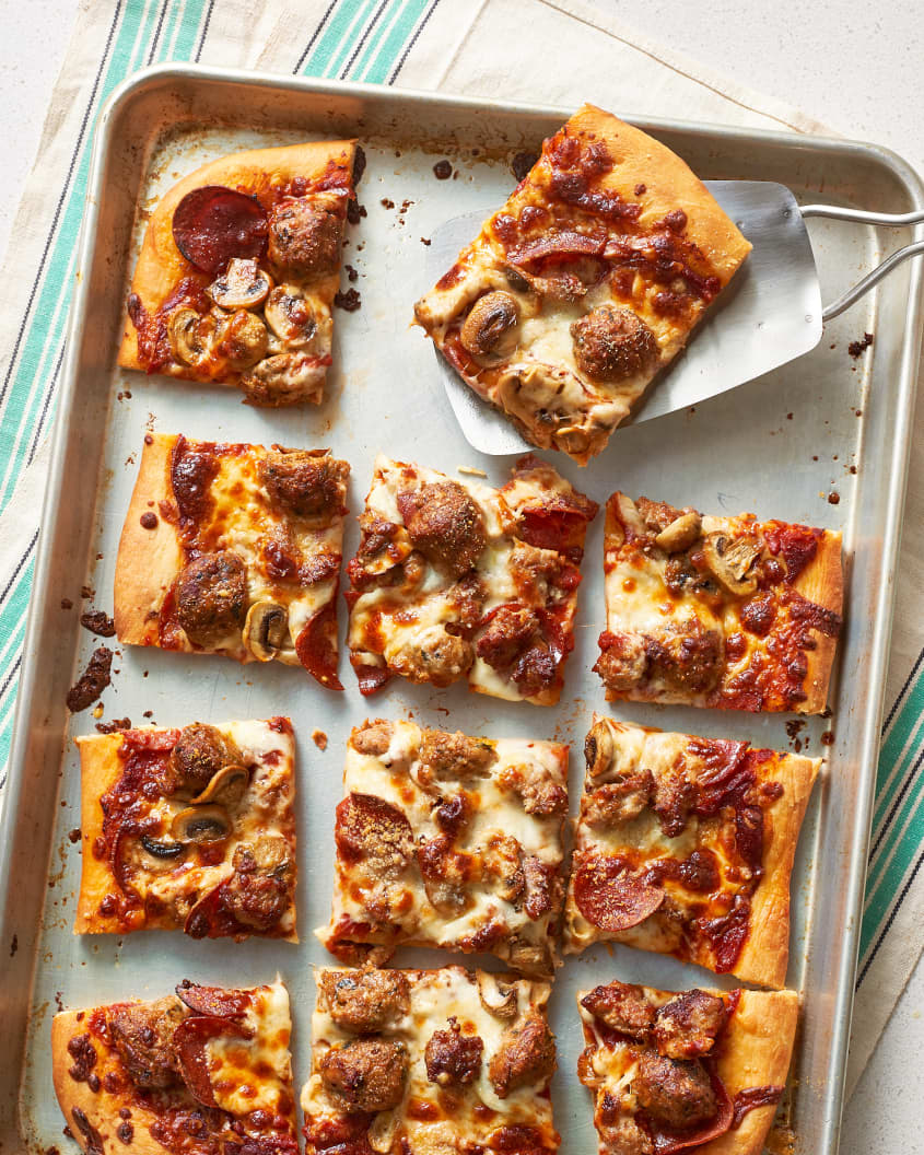 A slice of sheet pan pizza, topped with mushrooms, pepperoni, quartered meatballs, and pork sausage, is scooped out from a baking sheet using a metal spatula