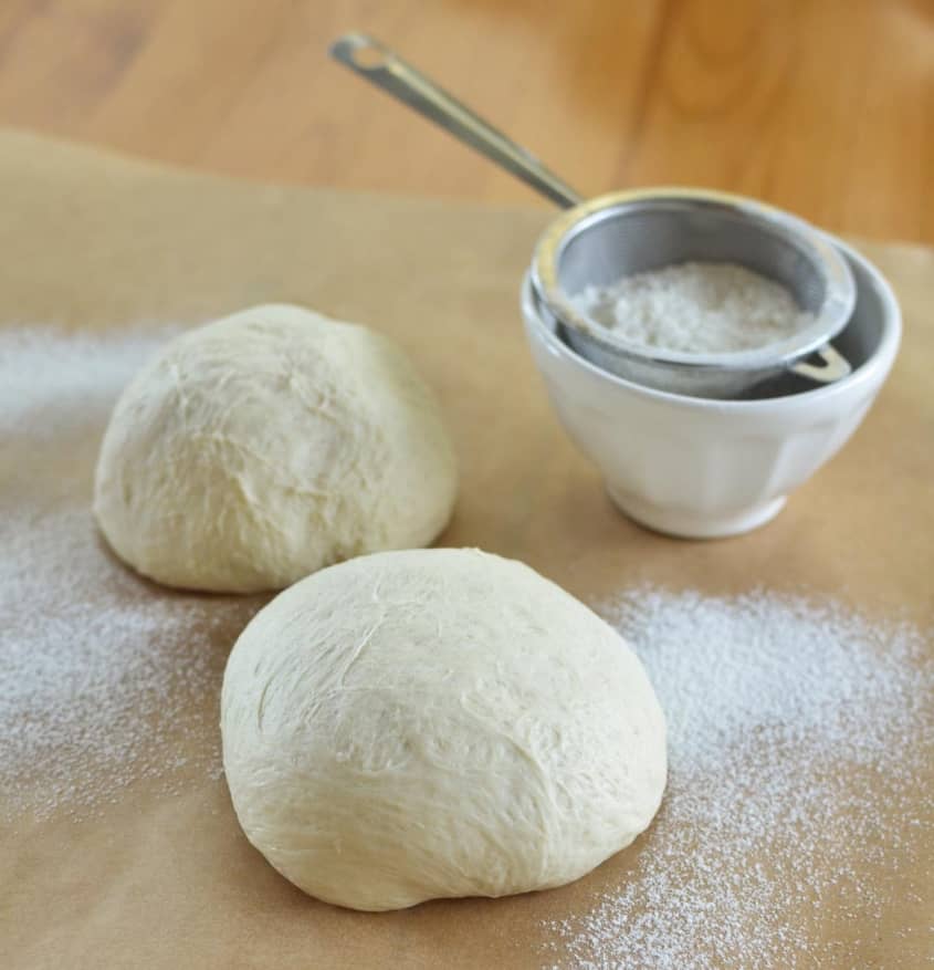 Two pizza dough, shaped in a circle, dusted with flour, with a seive over a small bowl on the side