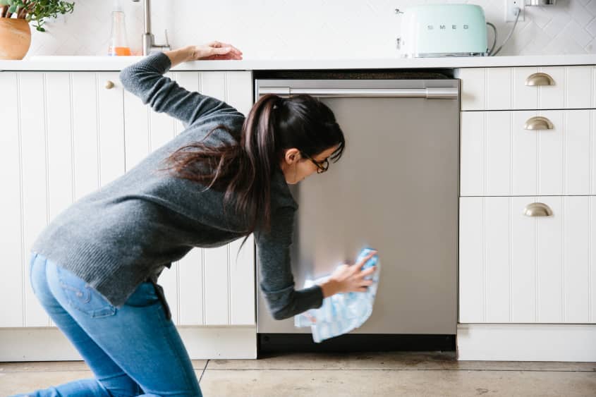 A woman is bent down wiping the exterior of a dishwasher