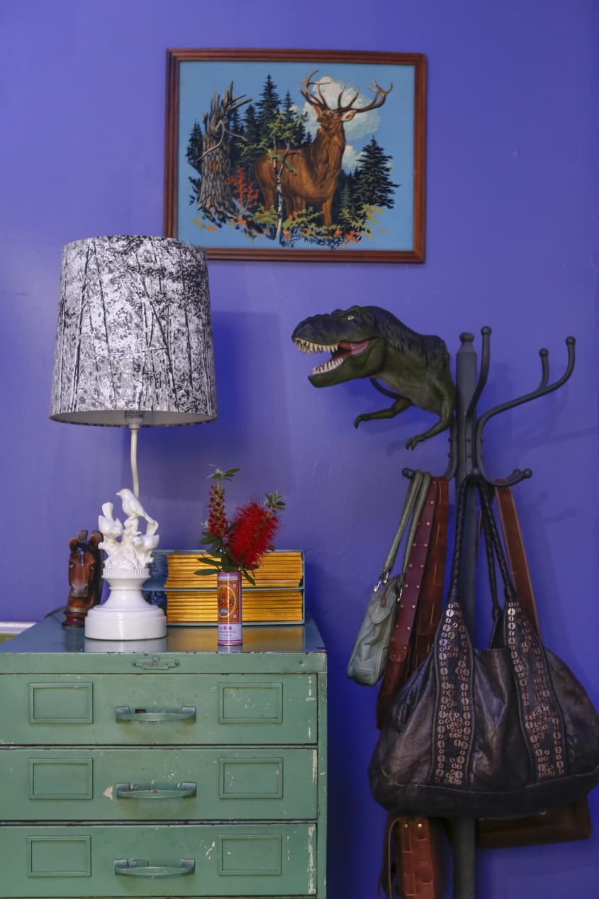 A purple wall in a living space; a green table has a lamp and tchotchkes on top. A coat rack holds purses and a dinosaur puppet