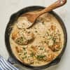 Chicken breasts in creamy sauce with herbs in a skillet, wooden spoon, and striped cloth.