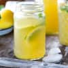 Mason jar of lemonade with lemon slices and mint, next to a carafe and ice cubes on a wooden table.