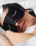 Woman sleeping with a black cat sleep mask and seal in upper left reads "Apartment Therapy The Best List 2024 Sleep Masks"