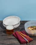 Two bowls of food, a bowl of rice, stacked speckled bowls, a glass of beer, and red napkins with chopsticks on a wooden table.