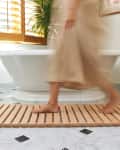 Person walking on a wooden bath mat in a bathroom with a white tub, marble floor, and wooden shutters.