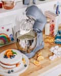 Blue stand mixer with whipped cream, cake with oranges and blueberries, eggs, and citrus slices on a wooden countertop.