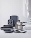 Cookware set with baking trays, muffin pan, saucepan, and utensils next to folded towels and a pillow on a white surface.