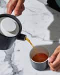 Pouring tea from a black teapot into a cup on a marble surface, with a Firebelly Tea box nearby.