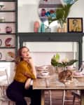 Woman in mustard blouse sitting at rustic table with pink dishes, surrounded by colorful plates and decor, cat on table.