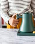 Person using a green electric kettle on a marble countertop, wearing a striped shirt and yellow skirt.