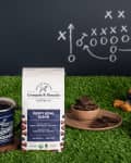Grounds & Hounds Puppy Bowl Blend coffee with footballs, mug, and snacks on artificial grass, chalkboard play diagram in background.